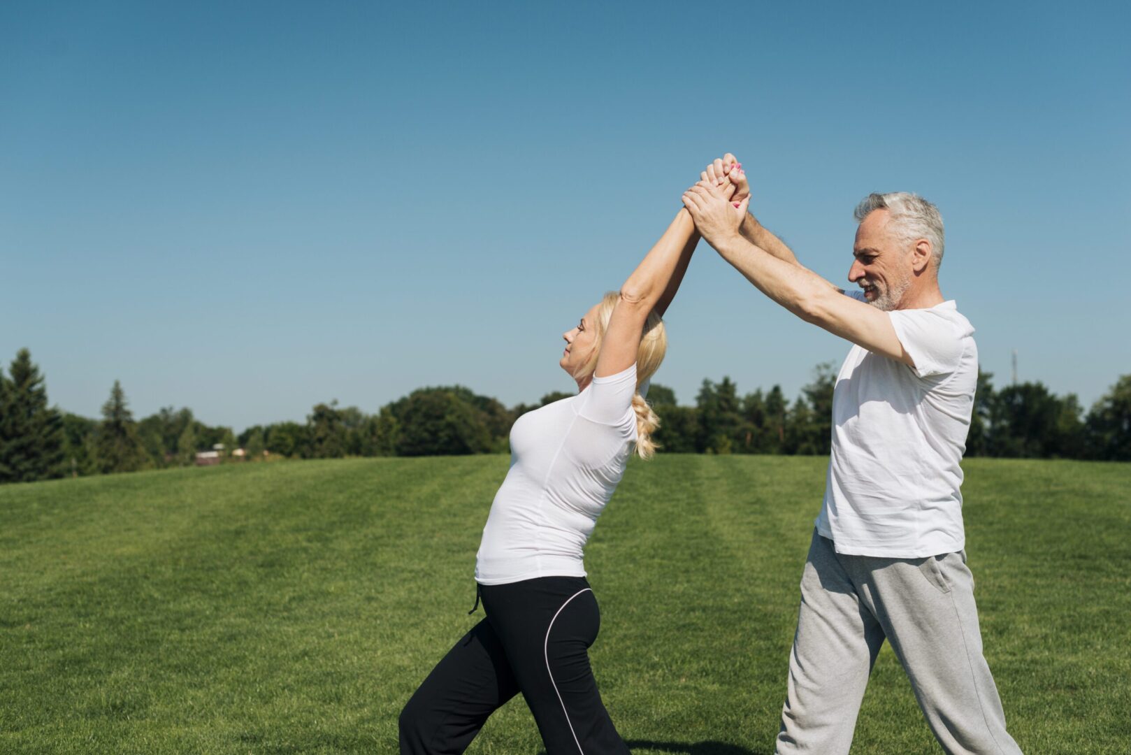 Pareja de personas mayores haciendo ejercicio al aire libre en un campo verde bajo un cielo despejado, estirando los brazos juntos, transmitiendo salud y bienestar.