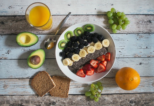 Tazón de desayuno saludable con frutas como fresas, moras, kiwi y plátano, acompañado de aguacates, jugo de naranja, uvas, pan integral y una naranja sobre una mesa rústica de madera.