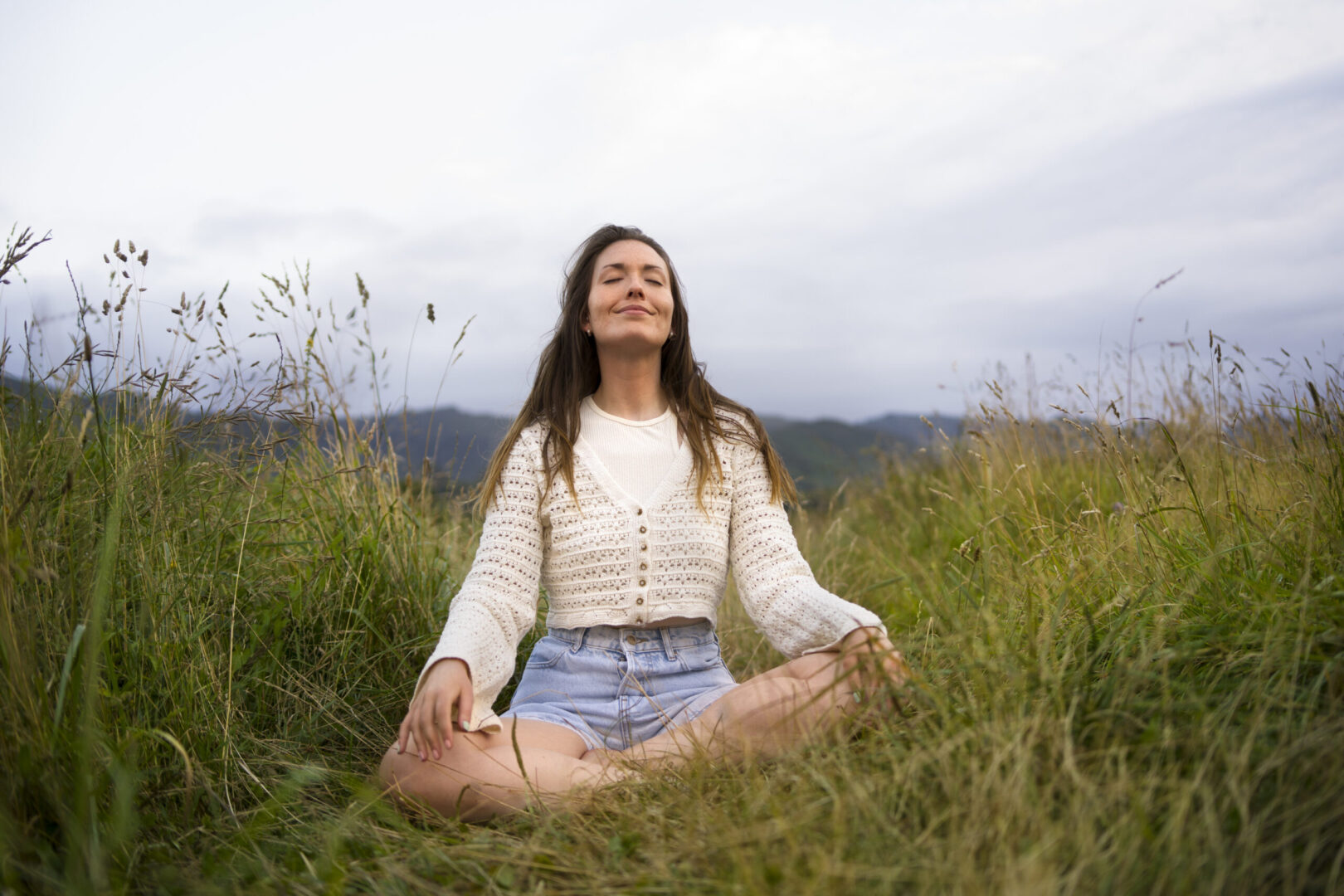 Mujer sentada en un prado, con las piernas cruzadas y los ojos cerrados, disfrutando de un momento de tranquilidad y conexión con la naturaleza.