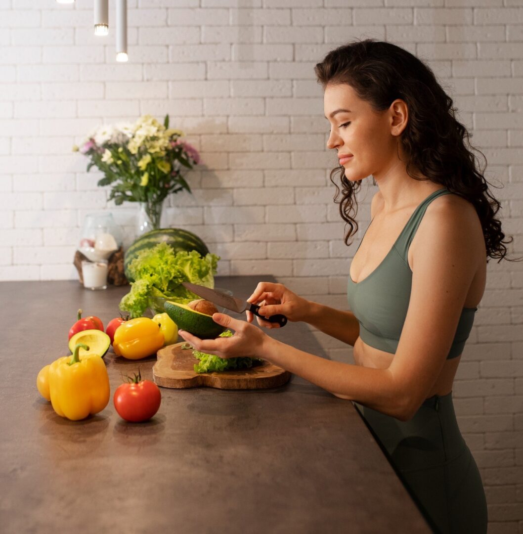 Una mujer en ropa deportiva corta un pepino en una cocina moderna, rodeada de vegetales frescos como pimientos y lechuga, sugiriendo un estilo de vida saludable.