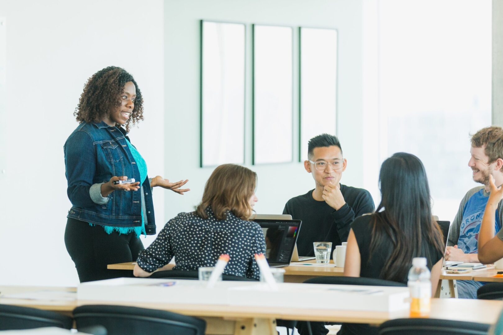 grupo de personas en una reunión de trabajo en una oficina moderna. Una mujer, de pie, parece estar liderando la discusión mientras los demás están sentados alrededor de una mesa, prestando atención. La escena refleja un entorno colaborativo y profesional