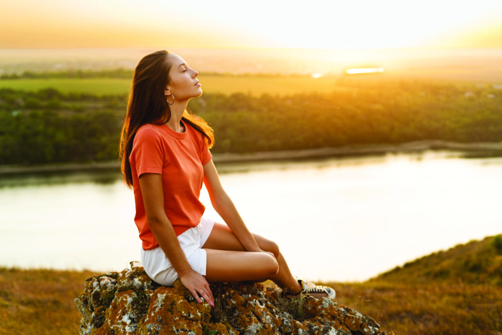 Mujer sentada en una roca al aire libre, disfrutando del atardecer junto a un río, con los ojos cerrados, transmitiendo tranquilidad y conexión con la naturaleza.